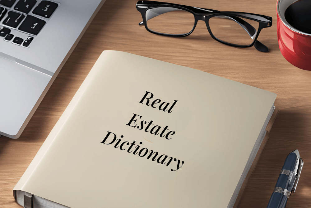 An image of a real estate dictionary on a table. Reading glasses and a laptop are present as well.